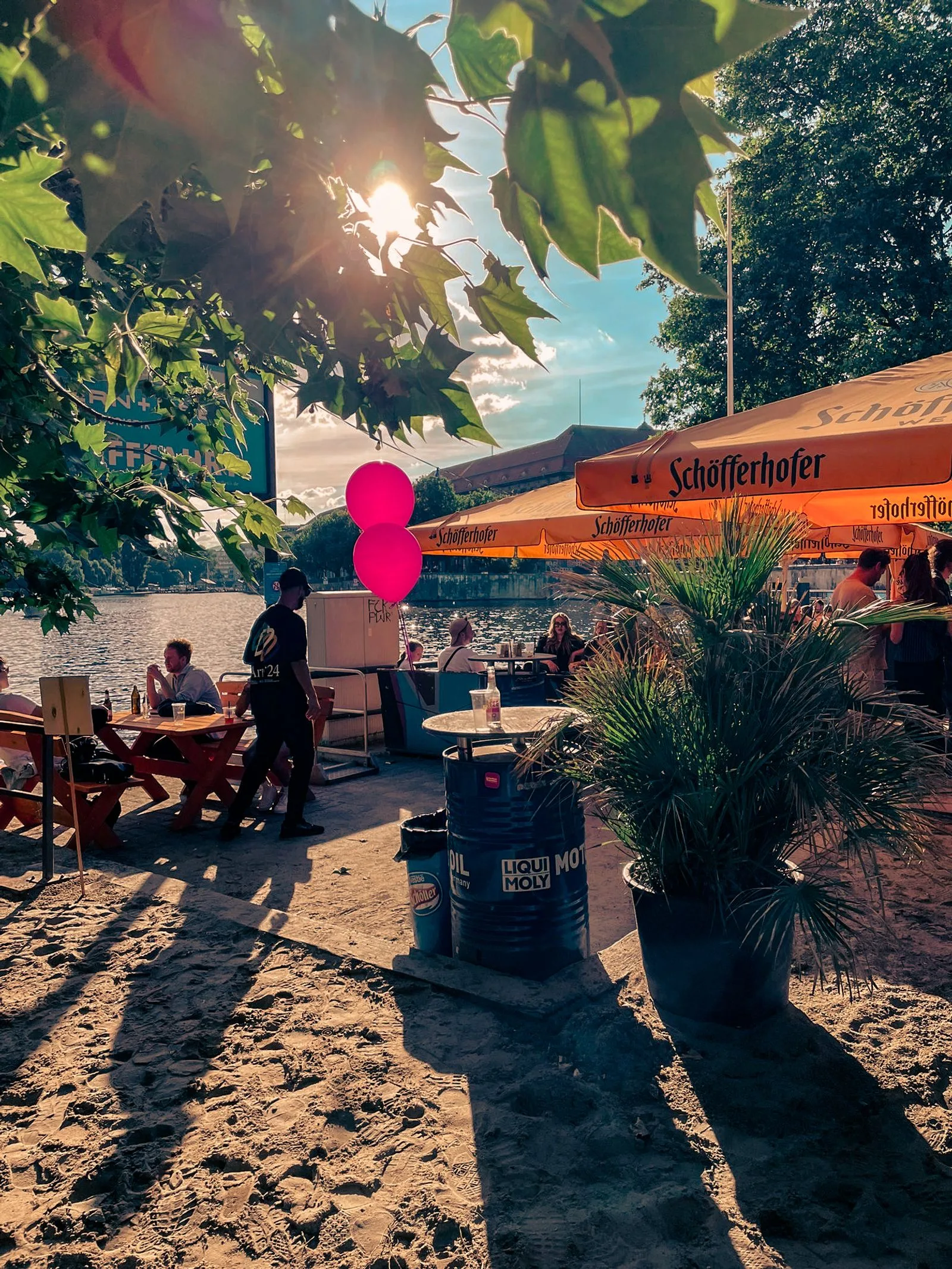 Sonniger Außenbereich des Gestrandet an der Spree mit orangenen Sonnenschirmen, Sitzplätzen im Sand und Gästen im Schatten.
